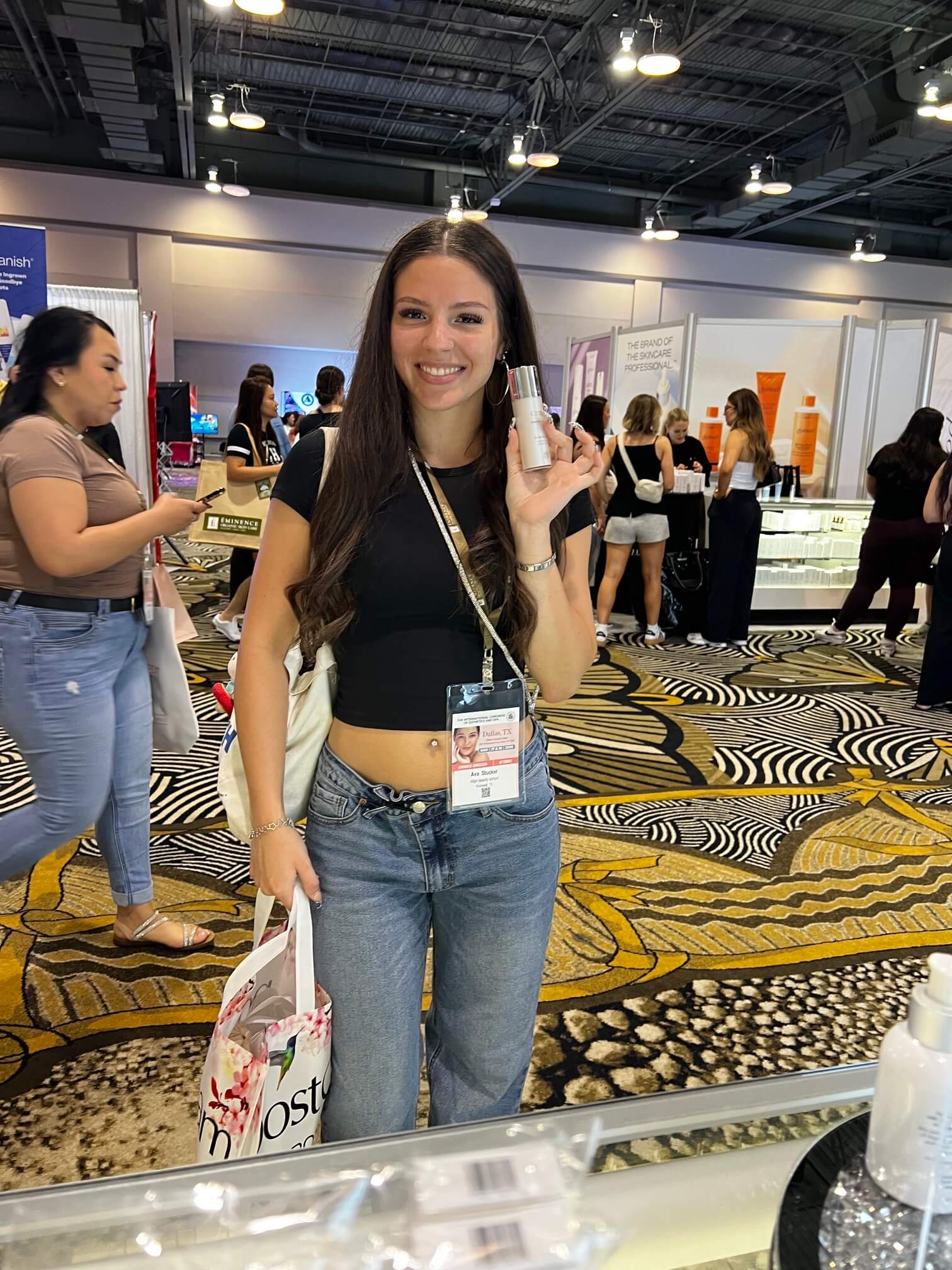 A smiling licensed esthetician holding an Enspri clinical skincare product at the ICES Dallas trade show booth.