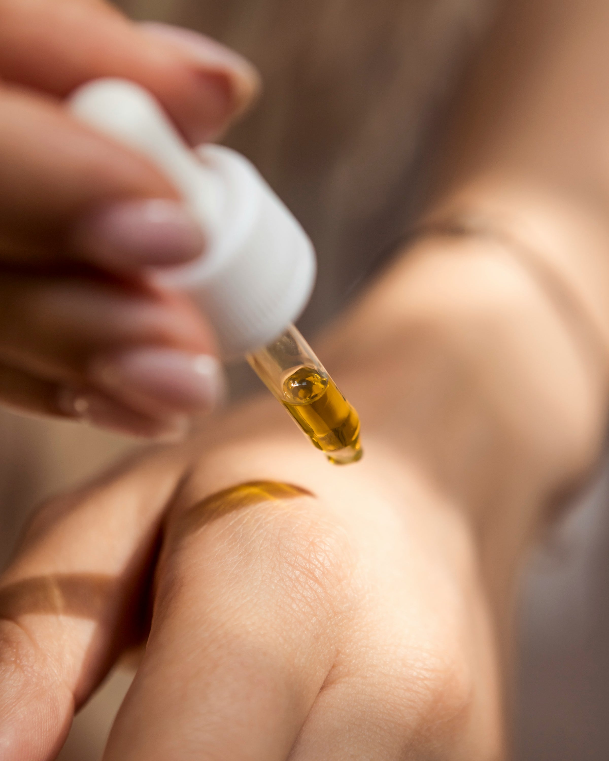 A close-up shot of a person using a glass dropper to apply a golden skincare oil onto the back of their hand.
